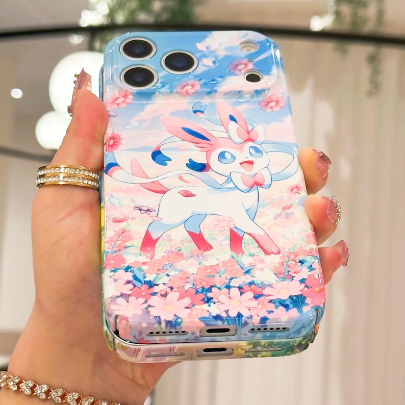 May include: A phone case with a cartoon Sylveon design. The case has a blue sky and floral background with pink and white flowers. The Sylveon is white, pink, and blue. The case has a glossy finish.