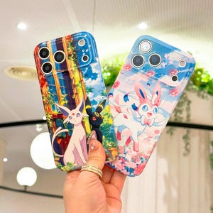 May include: Two phone cases with vibrant designs. One case displays a white and black cartoon character against a colourful backdrop. The other features a cartoon character with pink and blue, set against a floral and sky background.