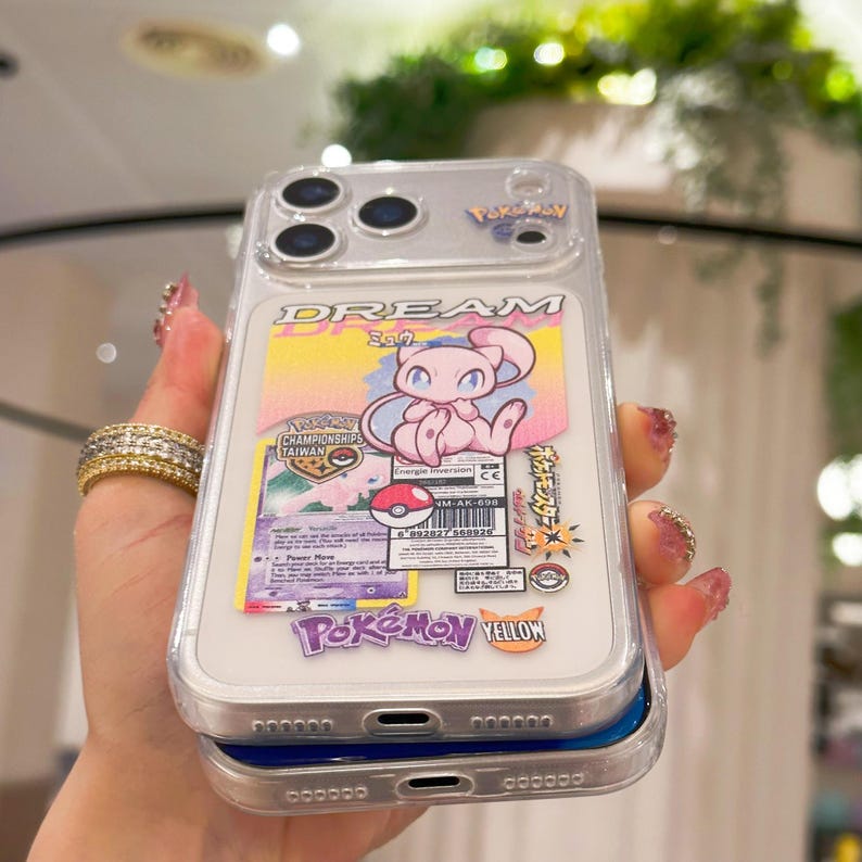 May include: Clear phone case featuring a pink and white Pokemon card design with the text "DREAM" and "Pokemon Yellow". The case has a clear border and a blue base. The design includes a cartoon character.