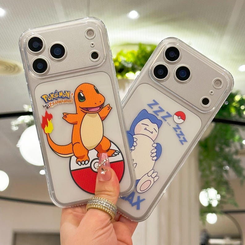 May include: Two clear phone cases with cartoon graphics. One features an orange Charmander character on a red and white Pok&eacute; Ball, the other a blue Snorlax with ZZZs. The cases have a clear border and are held by a hand.