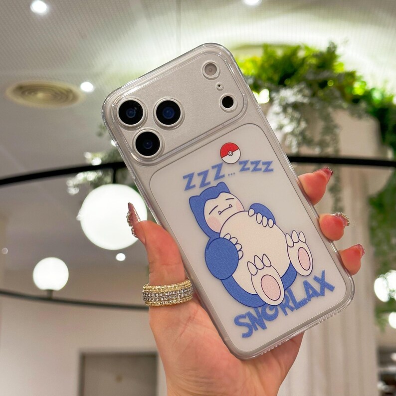 May include: Clear phone case featuring a blue Snorlax character sleeping, with the text "Zzz...Zzz" and "SNORLAX". The case has a clear border and a camera cutout.