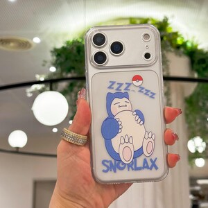 May include: Clear phone case featuring a cartoon Snorlax design. The design includes a blue Snorlax character with the text "Snorlax" and "Zzz...Zzz". The case has a silver camera module.