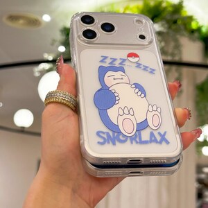 May include: Clear phone case featuring a blue Snorlax cartoon character with the word "SNORLAX" in blue. The design includes "ZZZ" and a Pok&eacute; Ball. The case has a clear border and covers the phone's camera.