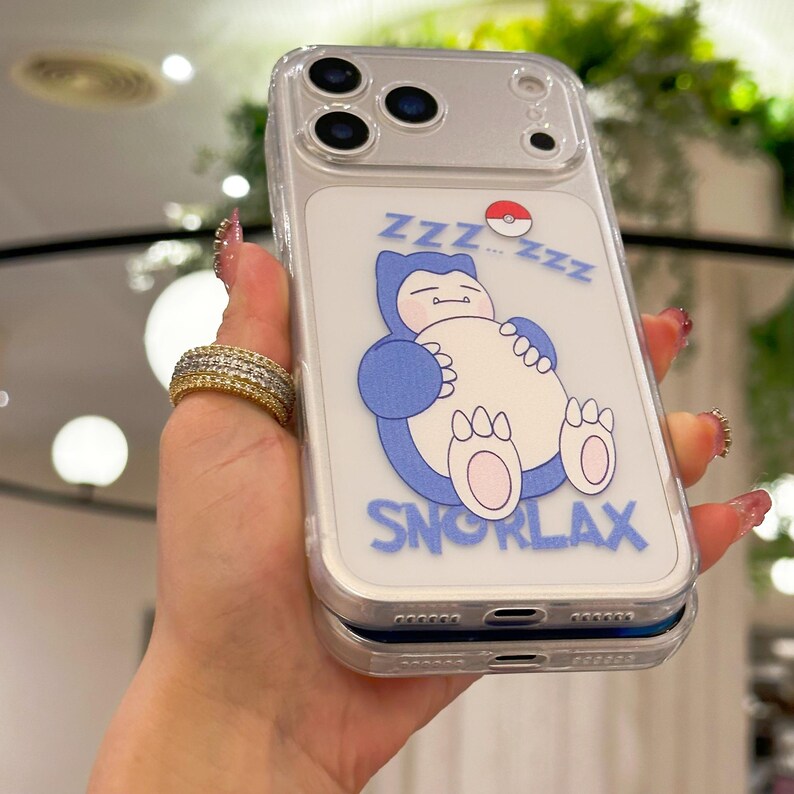 May include: Clear phone case featuring a blue Snorlax cartoon character with the word "SNORLAX" in blue. The design includes "ZZZ" and a Pok&eacute; Ball. The case has a clear border and covers the phone's camera.