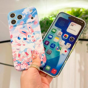 May include: A light blue phone case with a cartoon Sylveon character in pink and white, set against a sky and floral background. The case is held next to a phone displaying a home screen with app icons.