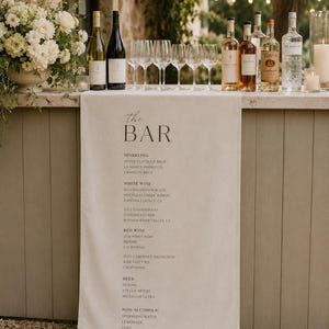May include: A bar display featuring bottles of wine and spirits, and wine glasses. A linen bar runner with the text "the BAR" lists sparkling, white, and red wines, beers, and non-alcoholic options. The setting is outdoors.