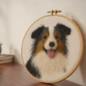 May include: A handcrafted dog portrait in an embroidery hoop. The dog's fur is black, brown, and white, with a pink tongue visible. The artwork is set against a white backdrop, framed in wood.