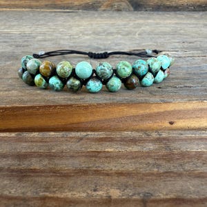 May include: Double-strand beaded bracelet featuring turquoise and brown beads. The beads are strung on a black cord, creating a layered effect. The bracelet is displayed on a wooden surface.