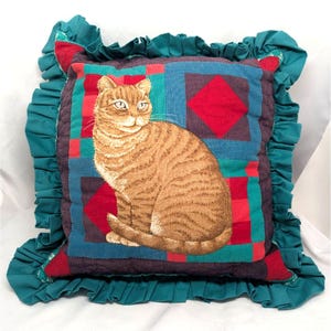 Puede incluir: Un cojín cuadrado decorativo con una imagen detallada de un gato atigrado. El cojín tiene un diseño de patchwork en rojo, azul y morado, con un borde de volantes verde azulado. El gato es naranja y marrón.
