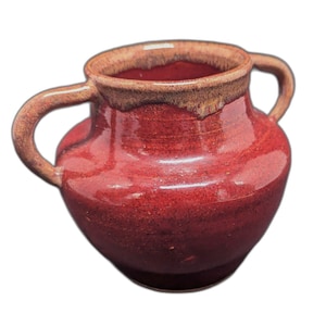 May include: A small, red ceramic vase with two curved handles. The vase has a rounded body and a slightly flared rim with a brown glaze. The overall design is rustic and handmade, suggesting a pottery piece.