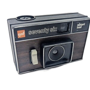 May include: A vintage GAF Seventy-Six instant load camera with a black and wood-grain finish. The camera has a lens, flash, and a silver switch. The text "gaf seventy six" is printed on the front.