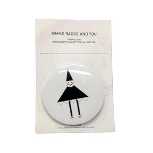 May include: A white button badge featuring a black and white cartoon figure with a triangular body and cone-shaped head. The badge is attached to a card with text: "MMMG BADGE AND YOU SPRING 2008 MMMG HAPPY MARKET SPECIAL EDITION".