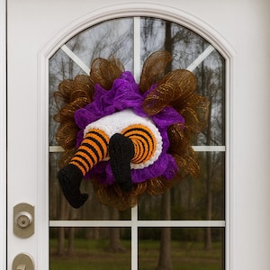 May include: A Halloween wreath featuring a whimsical design of a witch's legs and feet. The legs are striped black and orange, with black shoes. The wreath has a purple and brown mesh base, and is displayed on a white door.
