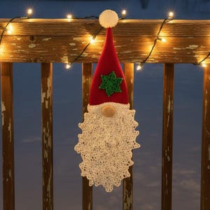 Puede incluir: Decoración de gnomo navideño hecha a mano. El gnomo tiene un gorro rojo con una estrella verde, una barba de ganchillo blanca y una nariz de madera. Cuelga de una barandilla de madera con luces de cadena.