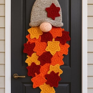 Könnte beinhalten: Handgefertigte gehäkelte Zwerg-Türkranz. Der Zwerg hat einen grauen Hut mit einem roten Stern, eine pfirsichfarbene Nase und einen Bart aus orangefarbenen, gelben und roten Blättern. Die Dekoration hängt an einer schwarzen Tür.