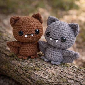 May include: Two crocheted bat plushies, one brown and one gray, with large black eyes and small white fangs. The bats have outstretched wings and are posed on a textured, brown log. The plushies are handmade.