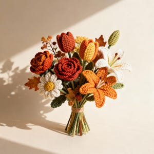 Op de afbeelding: Een handgemaakt gehaakt boeket met bloemen in herfsttinten. Het boeket bevat oranje lelies, rode rozen, gele en oranje tulpen, witte madeliefjes en witte lelies, samengebonden met touw.