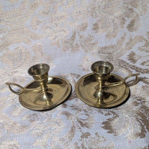 May include: A pair of polished brass candle holders, each featuring a wide circular base, a central cup for a taper candle, and a small looped handle attached to the side for easy carrying. The metal has a warm, reflective golden finish.