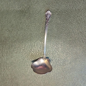 May include: A silver-toned metal ladle featuring a shell-shaped bowl and a slender handle with decorative scrollwork at the top and base. The polished surface reflects light, highlighting the curved contours of the bowl and the intricate detailing.
