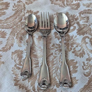 Puede incluir: Un juego de tres cubiertos de metal plateado que consta de una cuchara pequeña, un tenedor de cuatro dientes y una cuchara grande. Cada pieza presenta un diseño de mango con nervaduras verticales y un motivo decorativo de concha.