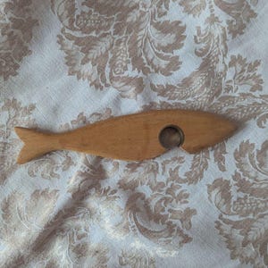 May include: A smooth, light brown wooden bottle opener shaped like a fish. The object features a carved, circular opening on the side, designed for removing bottle caps, with a simple, streamlined silhouette and a distinct tail fin.