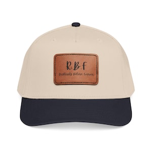May include: A beige baseball cap with a dark navy blue brim and a top button. Centered on the front is a rectangular brown leather patch featuring the black text R.B.F and the phrase Recklessly Believe Forever in a cursive script font.