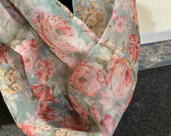 May include: A floral infinity scarf featuring a delicate pattern of pink and red roses, with yellow and green accents, set against a soft, pale blue background. The scarf is made of a lightweight, sheer fabric.