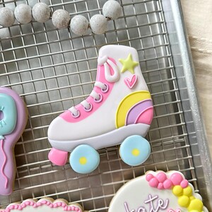 May include: A decorated sugar cookie in the shape of a roller skate. The skate is white with pink accents, a yellow star, a pink heart, and a rainbow design. The wheels are light blue with yellow centers. The cookie is on a metal cooling rack.