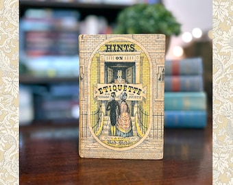 Peut inclure: Un livre vintage intitulé "Hints on Etiquette and Usages of the Society" avec une illustration d'un couple devant un bâtiment. Le livre a une couverture beige avec des détails noirs et jaunes. L'arrière-plan comprend une bibliothèque floue et d'autres livres.