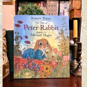 May include: A hardcover book titled The Tale of Peter Rabbit by Beatrix Potter, illustrated by Michael Hague. The cover features a brown rabbit in a blue jacket peering over a red wooden fence, surrounded by colorful flowers and green foliage.