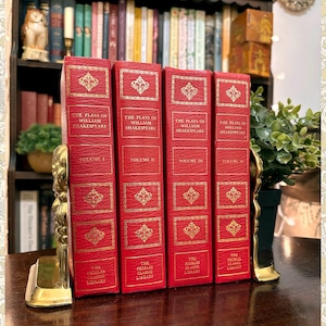 May include: Four red books titled "The Plays of William Shakespeare" with gold lettering and decorative accents. The books are held by gold bookends. The books are part of "The Priedler Classic Library" series.