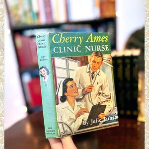 May include: Vintage book titled "Cherry Ames Clinic Nurse" by Julie Tatham. The cover features a retro illustration of a nurse and a doctor. The book has a green spine with the title in yellow and white lettering. The book is held in front of a blurred background of other books.