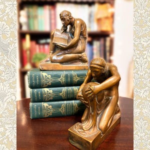 May include: Two bronze-toned bookends featuring kneeling figures, one reading an open book and the other holding a sphere, positioned on a stack of three green books titled Tolstoy's Works with gold decorative spine detailing.