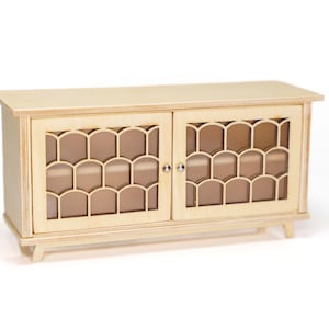 May include: A light-colored wooden sideboard featuring two cabinet doors with a decorative scalloped lattice overlay. The piece stands on four short, tapered legs and includes small, round silver-toned knobs on each door for opening.