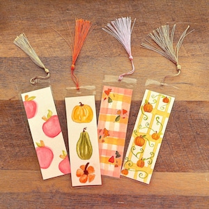 May include: Four rectangular bookmarks with clear protective sleeves and decorative tassels. The first features pink apples, the second shows a yellow pumpkin and green gourd, the third has a plaid pattern with leaves, and the fourth displays pumpkins on a trellis.