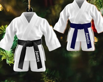 May include: Two white karate uniform ornaments with black and navy blue belts. One ornament has the name "James" on the belt, and the other has "Amelia." Both ornaments are hanging from gold strings, perfect for holiday decor.