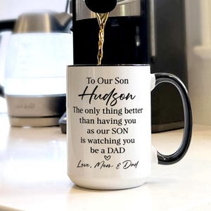 May include: White ceramic mug with a black handle and rim. The mug has the text "To Our Son Hudson" and a message about being a dad. Coffee is being poured into the mug.
