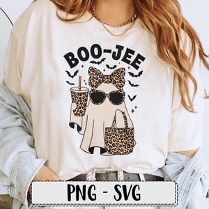 May include: Cream-colored t-shirt with a Halloween-themed graphic. The design features a ghost wearing sunglasses and a leopard print bow, holding a coffee cup and a tote bag. The text "BOO-JEE" is above the ghost, with bats and stars.