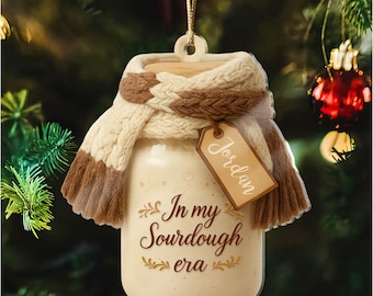 May include: A cream-colored, jar-shaped Christmas ornament with a wooden lid and a knitted scarf in cream and brown. The ornament has the text "In my Sourdough era" and a tag that says "Jordan".