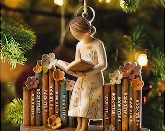 May include: A Christmas ornament featuring a wooden figure of a person reading a book, with the name "Sophia" at the base. The figure is surrounded by a fence with the text "BOOK TITLE 1" on each post, and adorned with flowers. The ornament hangs from a gold string.