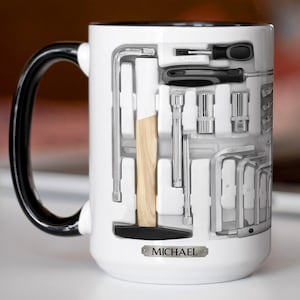 May include: White ceramic mug with a black handle and rim, featuring a toolbox design. The design includes a hammer, screwdrivers, and wrenches. The name "MICHAEL" is printed on a small metal plate on the mug.