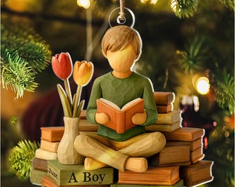 May include: Wooden ornament featuring a figure reading a book, seated on a stack of books. A vase with tulips is to the left. The text on the books reads "Just Who Loves A Boy Books Lucas". The ornament is suspended by a gold string.
