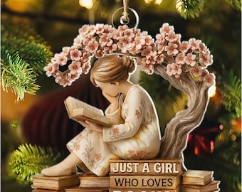May include: A decorative ornament featuring a girl reading a book under a flowering tree. The ornament reads "Just a girl who loves books" and "Emily". The ornament is made of a light-colored material with a gold string for hanging.