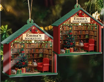 May include: Two Christmas ornaments shaped like miniature bookstores. Each ornament features a green roof, red walls, and a sign that reads "Emma's Bookstore." Inside, a Christmas tree, a red armchair, and bookshelves are visible. The ornaments are suspended by gold strings.