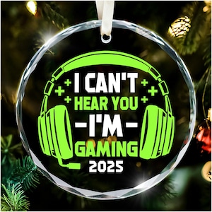 May include: A clear, round Christmas ornament with a green graphic of headphones and the text "I CAN'T HEAR YOU - I'M GAMING 2025". The ornament has a white ribbon for hanging.