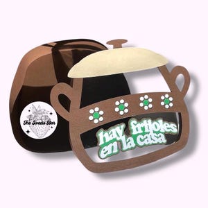 May include: A brown paper cutout of a pot with a light tan lid and handles. The pot has white and green flower accents and the text "hay frijoles en la casa" in green. A brown paper container is behind the pot.