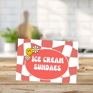 May include: A rectangular sign with a red and white checkered pattern and the words "ICE CREAM SUNDAES" in white. A smiling yellow face and white daisies are on the left side of the sign. The sign is on a wooden surface.