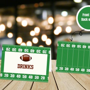 Pode incluir: Cartões de mesa com tema de futebol com um design de campo verde e linhas de jardas. Um cartão exibe uma bola de futebol e a palavra "DRINKS". O outro cartão mostra a parte de trás do cartão com o design do campo.