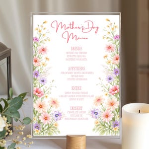 May include: A clear acrylic Mother's Day menu with floral watercolor accents. The menu features a pink script title and a list of drinks, appetizers, entrees, and desserts. The floral border includes pink, purple, and yellow flowers.