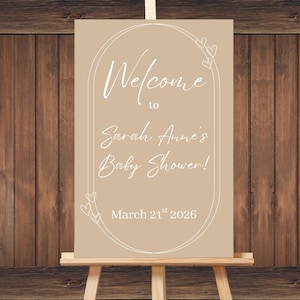May include: A beige baby shower sign on an easel with the text "Welcome to Sarah Anne's Baby Shower!" in white script. The date "March 21st 2026" is also displayed. The background is a wooden wall.